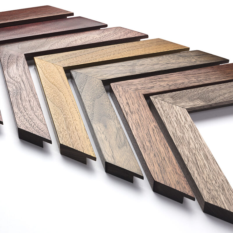 Eight wooden picture frame corners in various wood finishes are arranged in a zigzag pattern on a white surface, showcasing versatile styles that could complement anything from a floating frame for canvas to an elegant football shirt frame.