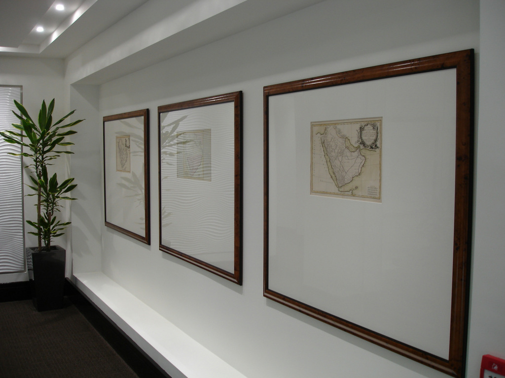 Three framed vintage maps hang on a white wall, much like how a football shirt frame might proudly display cherished memories. A potted plant is on the left side, and the ceiling features recessed lighting, casting a warm glow over the scene.