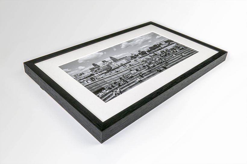 Framed black and white photograph lying flat in a circle frame, depicting a cityscape with buildings and people, against a plain white background. This piece stands out among art prints for its captivating simplicity, offering a timeless view that captures the essence of urban life.