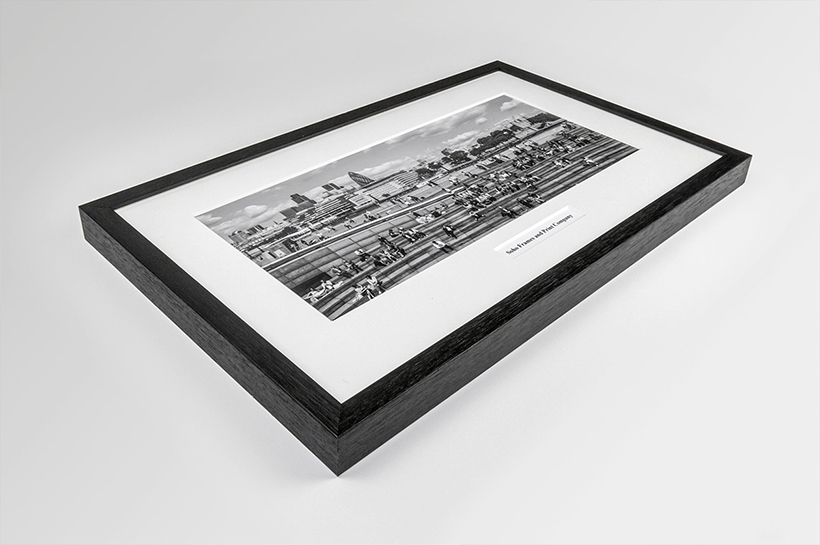 A framed black and white photograph of a cityscape with people elegantly complements an array of art prints. The dark-colored frame rests gracefully on a light gray surface, echoing the sophistication of a carefully curated football shirt frame display.