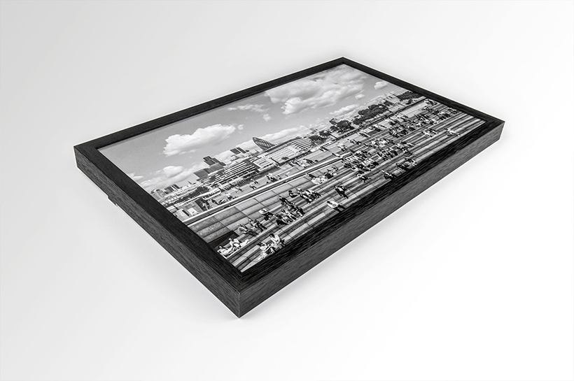 Framed black and white aerial photograph art print of a bustling city walkway with people, buildings, and clouds in the background, viewed from an angled perspective.