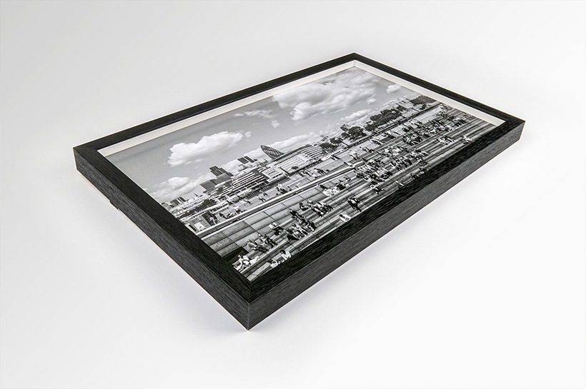 Framed black and white photograph of a cityscape with people on a rooftop, displayed alongside art prints on a pristine white surface.