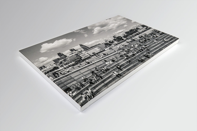 A black and white photograph features people sitting on wide, tiered steps in an urban area, casually observed through a circle frame. City buildings loom in the backdrop under a cloudy sky, adding depth to this candid moment amidst the hustle and bustle.