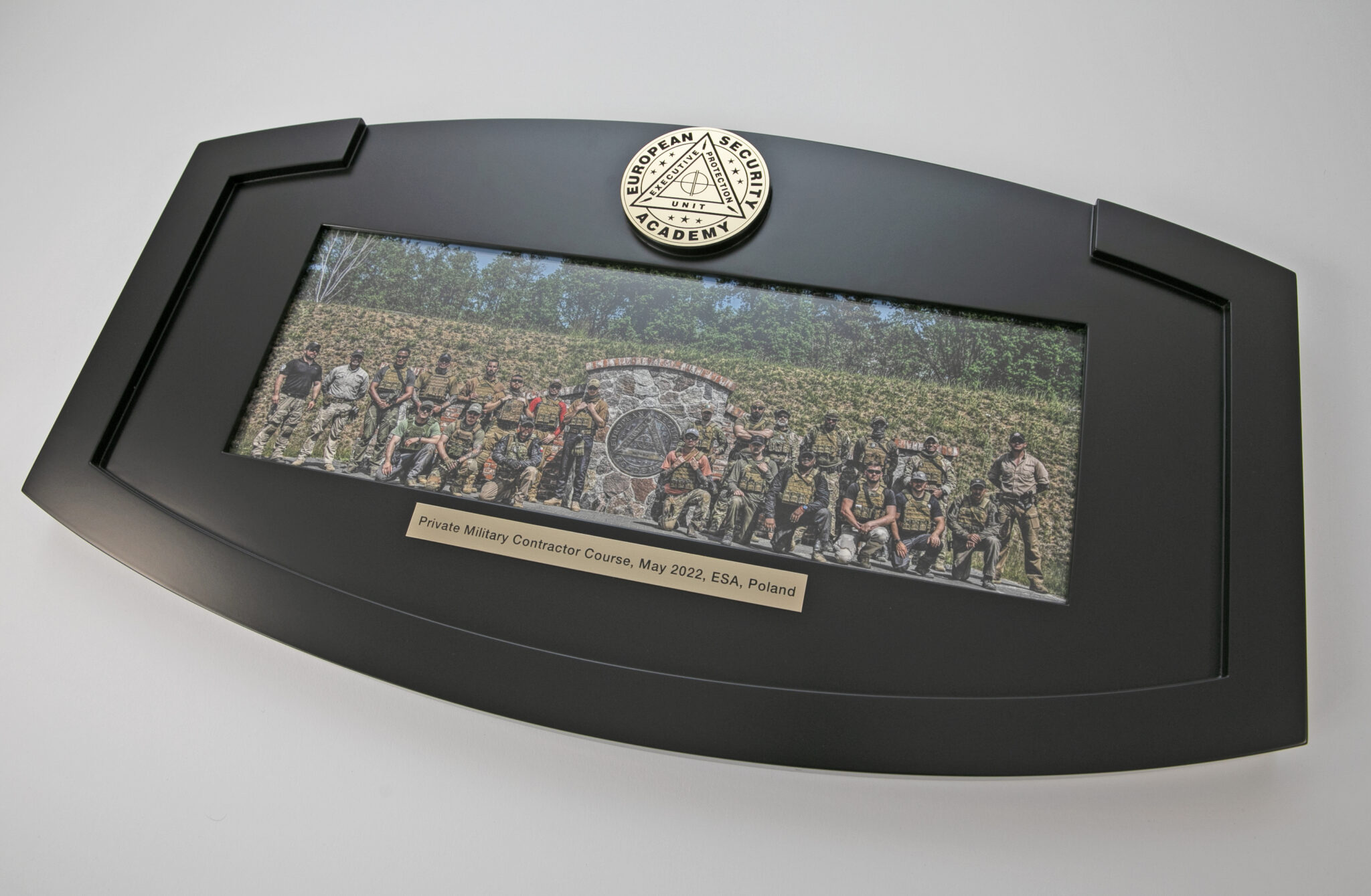 Framed panoramic photo of a group in military attire, encased in a distinctive black frame. The brass plaque reads, "Private Military Contractor Course, May 2022, ESA, Poland," and features the "European Security Academy" emblem engraved into brass on top.