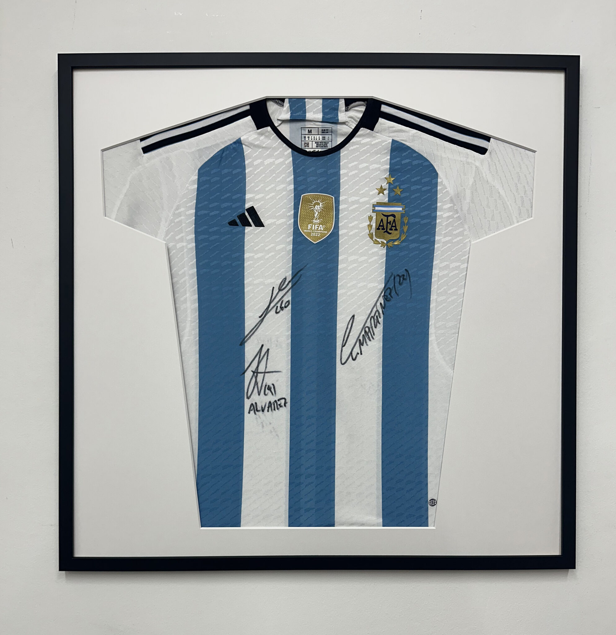 Framed Argentina soccer jersey with blue stripes and signatures displayed against a white wall.
