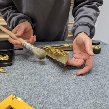 Person applies gold leaf to a picture frame with a brush on a gray work surface.