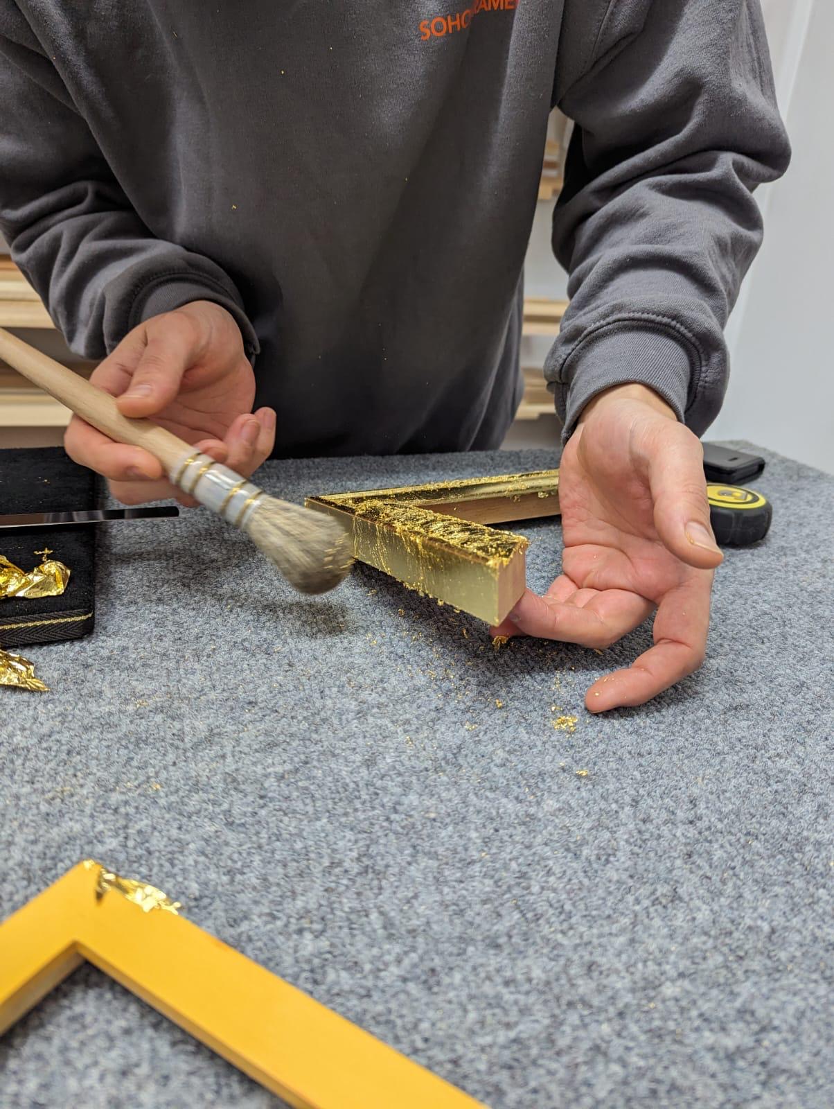 Person applies gold leaf to a picture frame with a brush on a gray work surface.