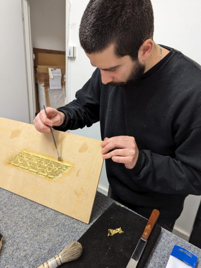 A man applies gold leaf to a rectangular pattern on a wooden board using a small brush.