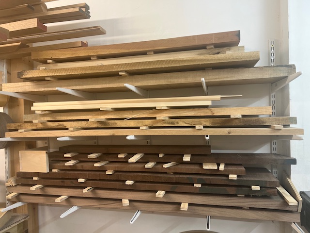 Stacks of various wooden boards organized on horizontal wall-mounted shelves in a workshop.