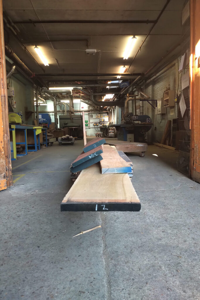 Long wooden planks and metal pieces are laid out on the floor inside an industrial workshop.