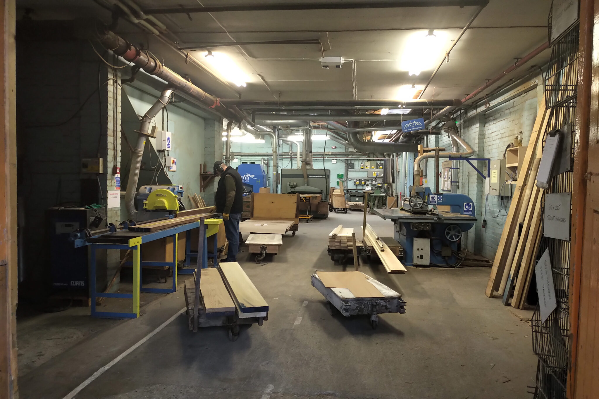 A person works in a woodshop with various tools, carts, and wooden planks on the floor.