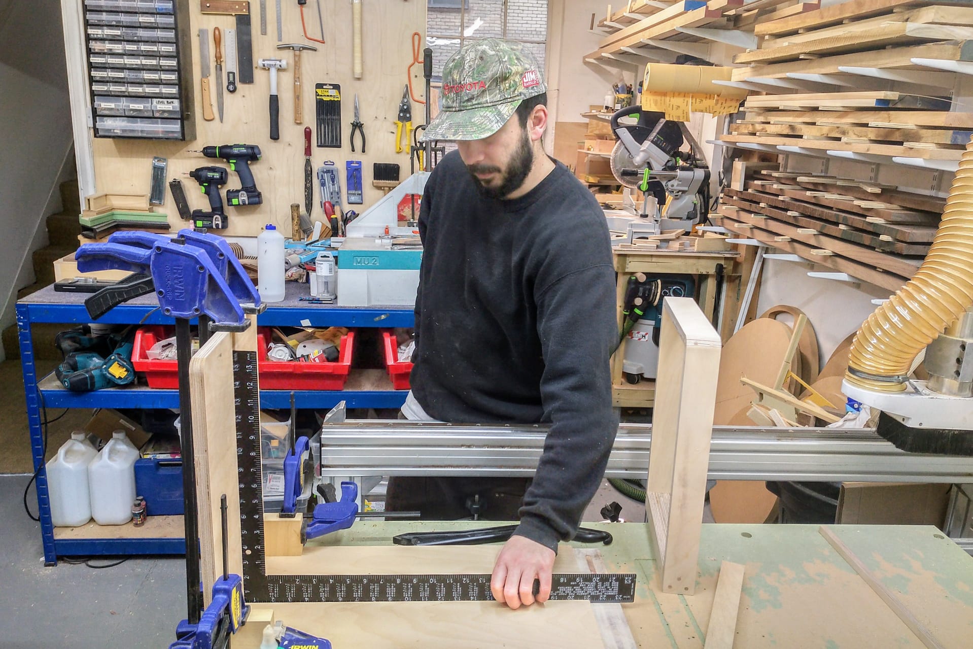 Man working in a woodshop, using clamps and a ruler to measure and assemble a wooden table.