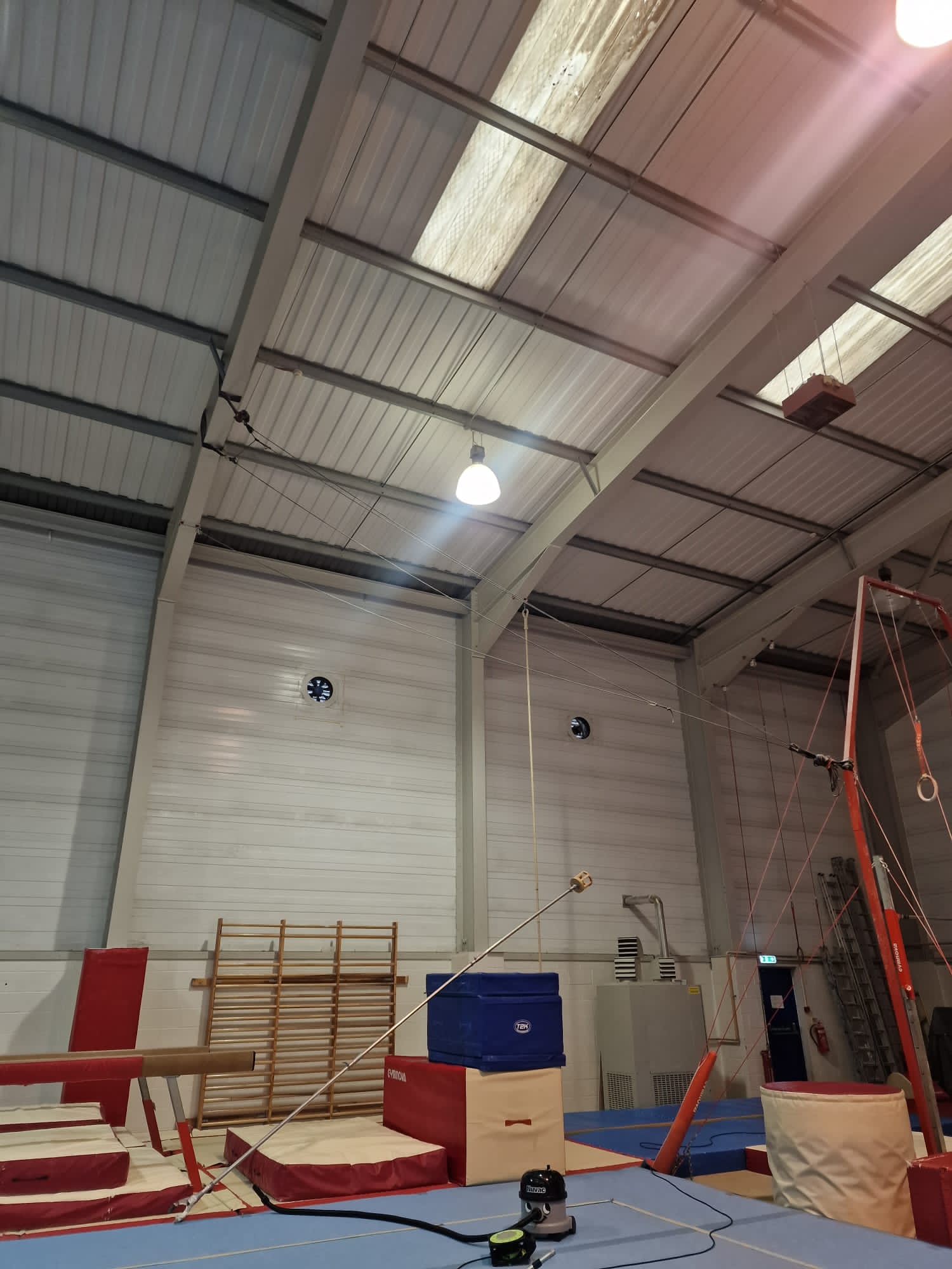 Indoor gymnastics training area with mats, equipment, and a beam under a high ceiling.