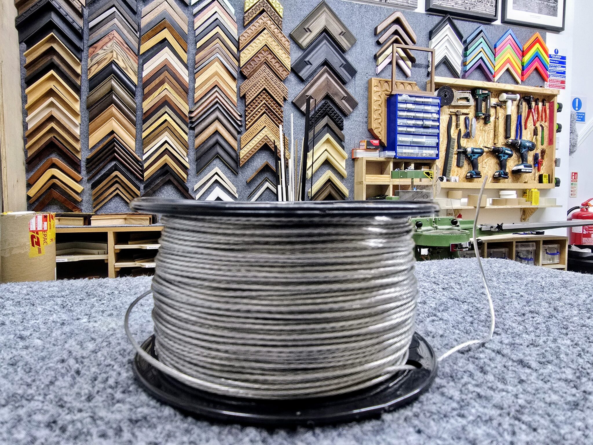 A spool of wire sits on a counter in a workshop with picture frames and tools on the wall.