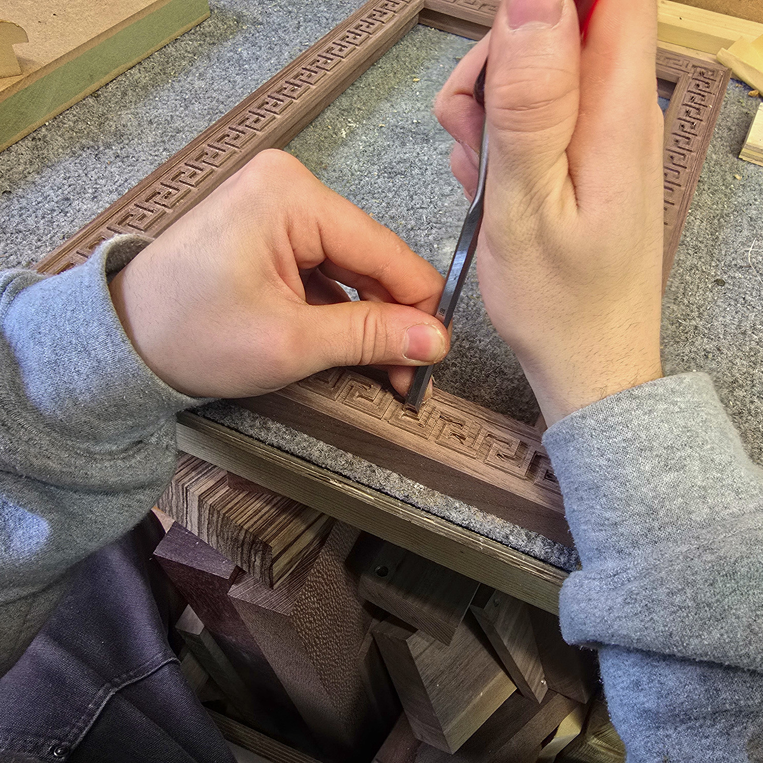 Hands carving a geometric pattern into a wooden picture frame rim using a chisel on a workbench.