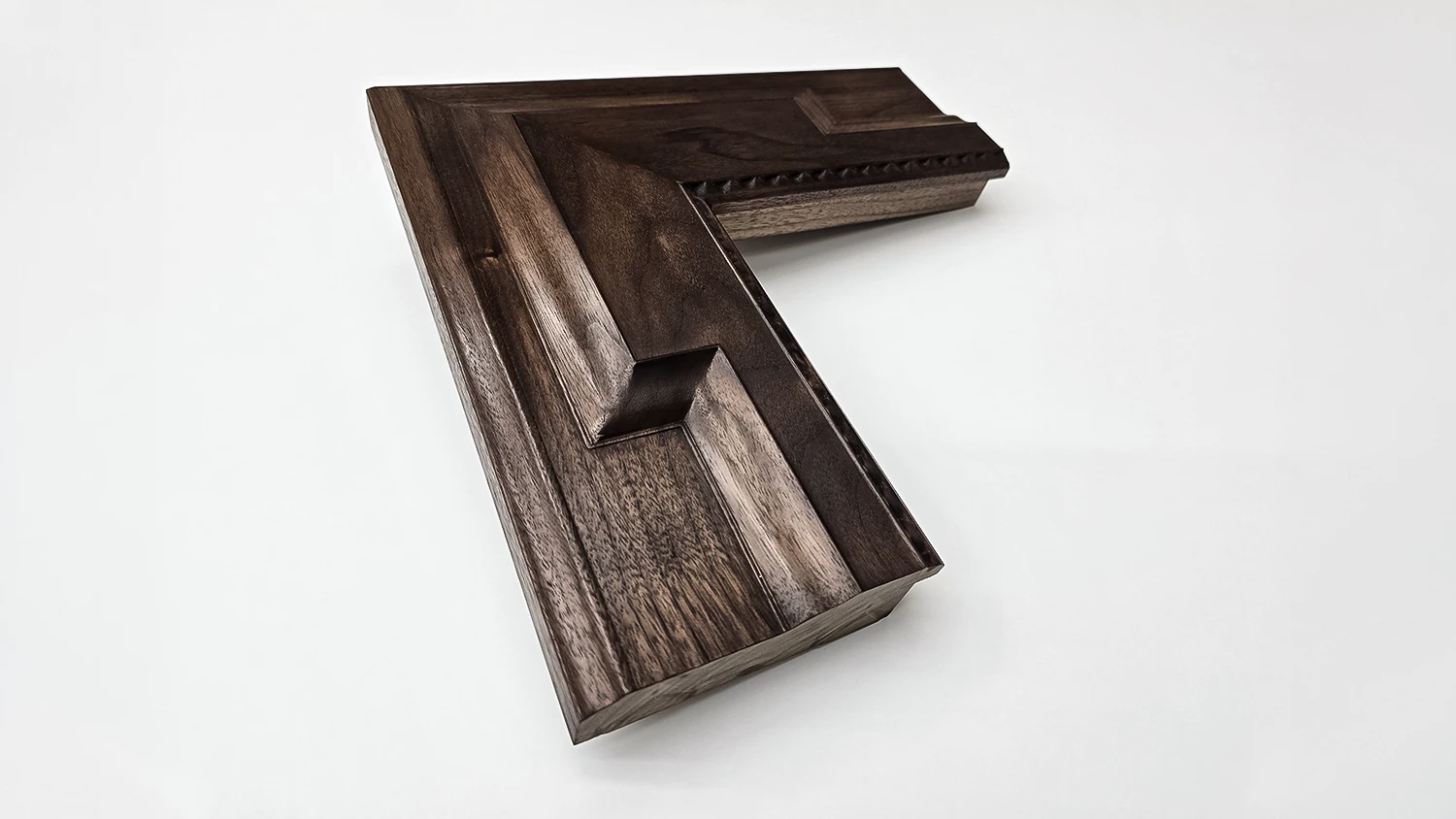 A wide picture frame corner in rich walnut wood with a carved bevelled face on a white background